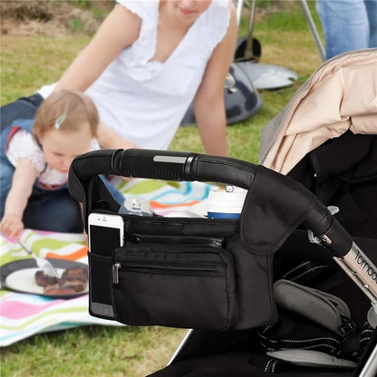 Black stroller organizer with phone and snacks attached to a stroller, with a child and adult in the background.