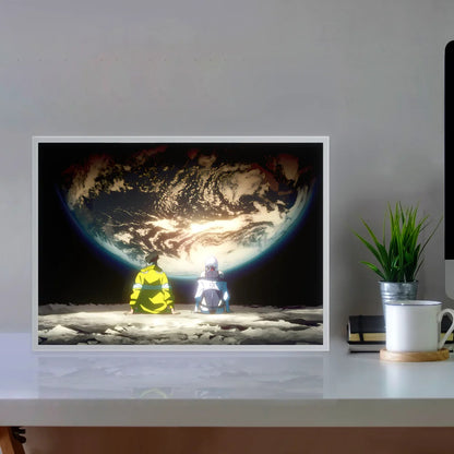 Framed artwork of two astronauts on the moon looking at Earth, placed on a desk with a plant and mug.